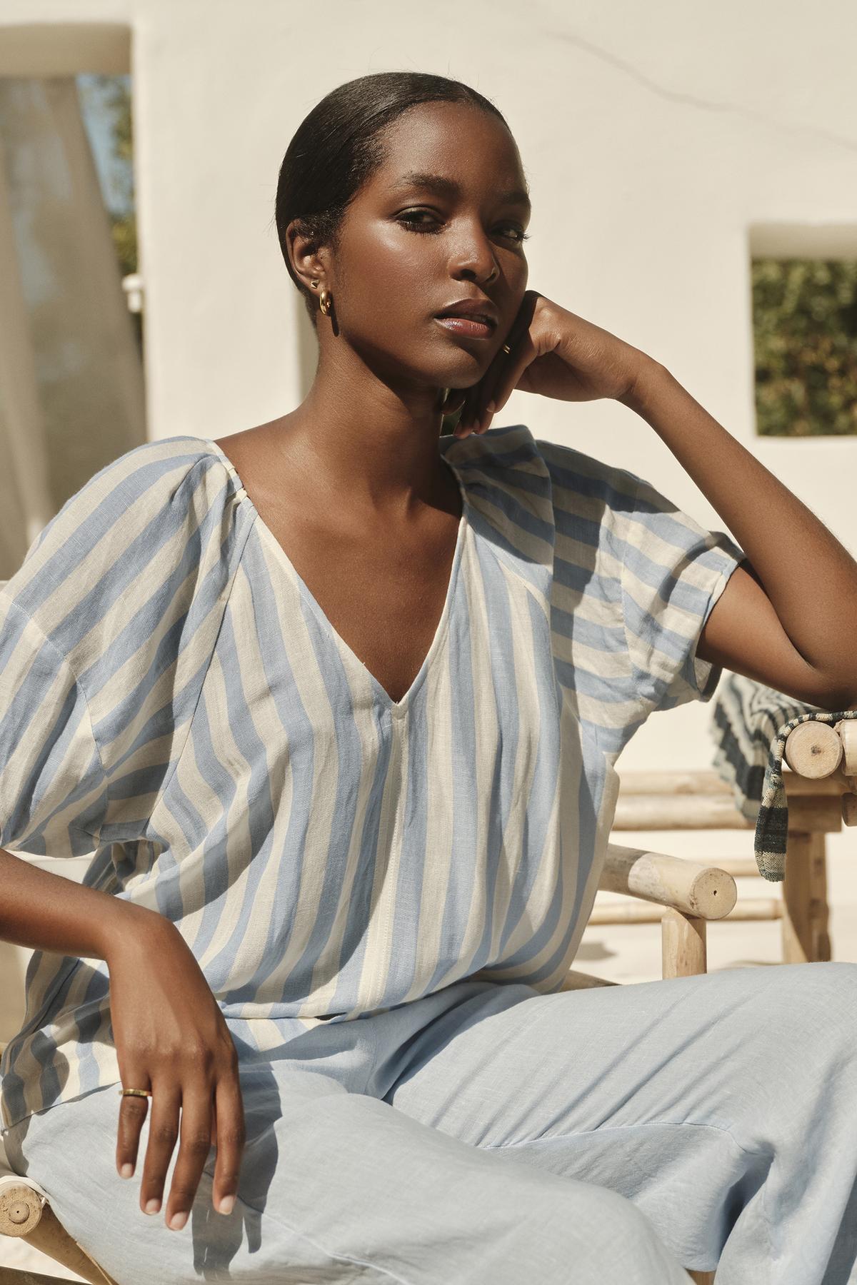 A woman in a KATY STRIPED LINEN TOP by Velvet by Graham & Spencer, with West Coast vibes, sitting on a chair.-36161171620033