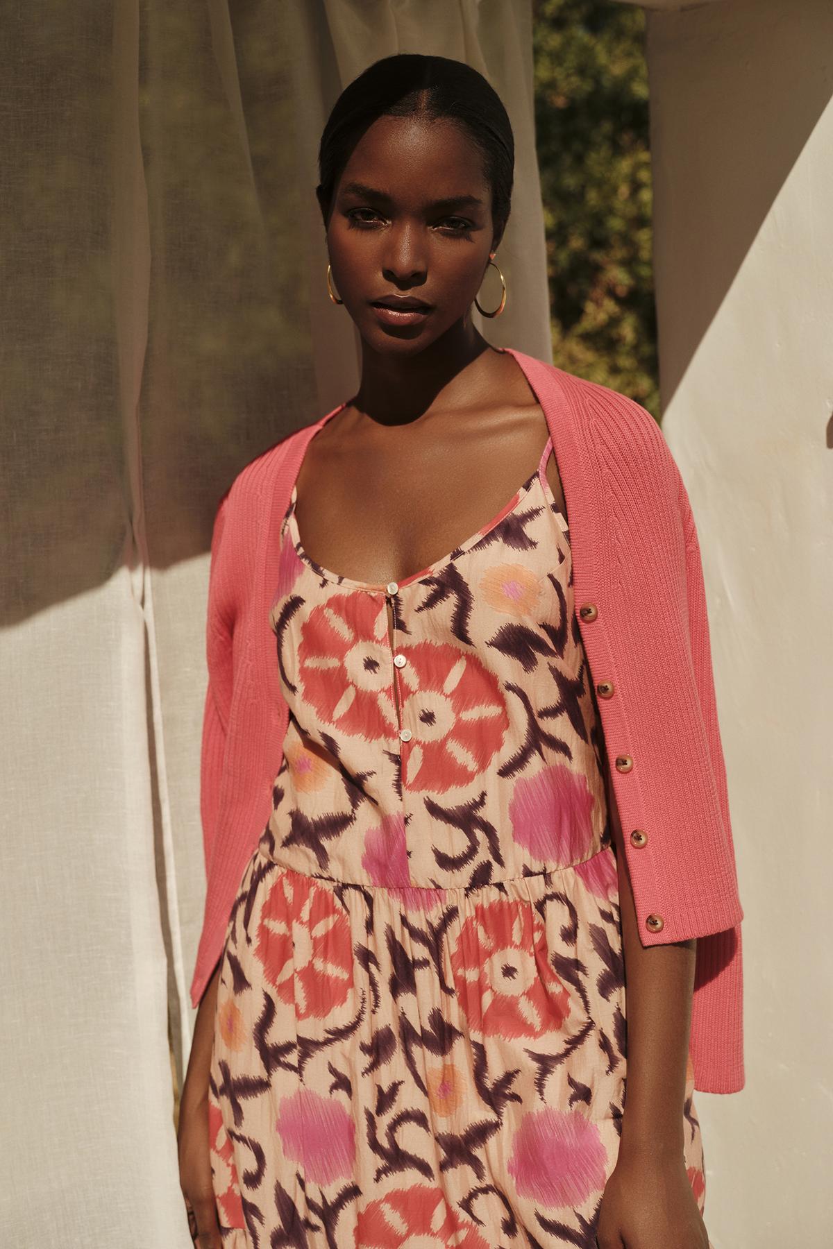   A woman in a floral dress and a Velvet by Graham & Spencer HYDIE BUTTON FRONT CARDIGAN stands confidently in sunlight, casting soft shadows around her. 