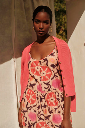A woman in a floral dress and a Velvet by Graham & Spencer HYDIE BUTTON FRONT CARDIGAN stands confidently in sunlight, casting soft shadows around her.