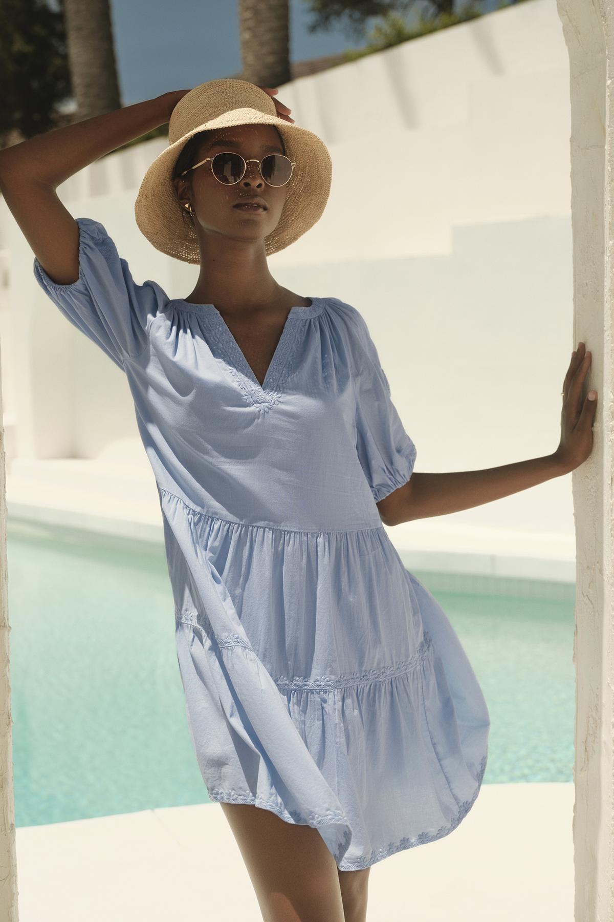   A woman in a CHRISSY EMBROIDERED BOHO DRESS by Velvet by Graham & Spencer and straw hat posing by a pool, with a white wall and palm trees in the background. 