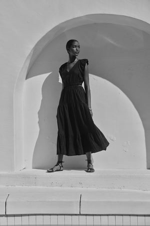 A woman in a flowing black GINGER LINEN CUT OUT TIERED DRESS by Velvet by Graham & Spencer stands elegantly against a white arched wall, casting a long shadow in the sunlight.