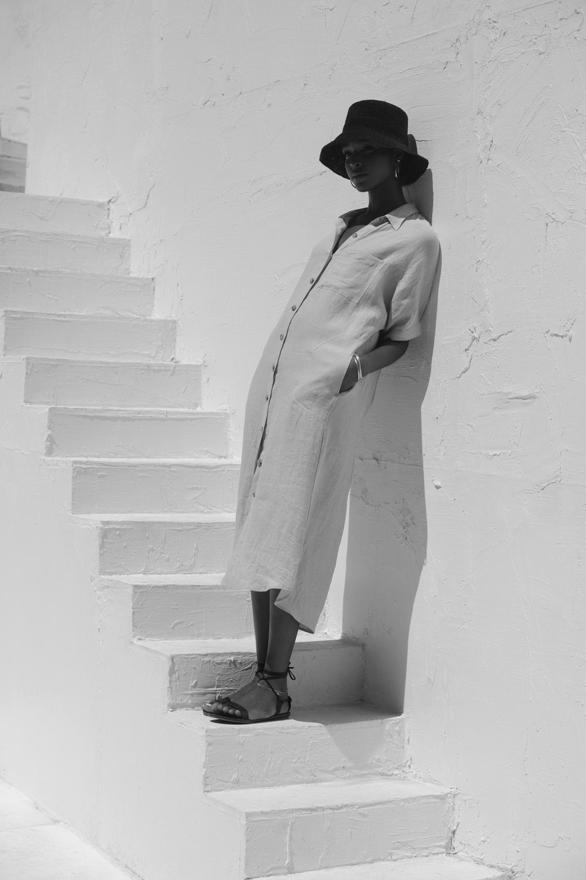   A person in a long SANDRA LINEN DRESS from Velvet by Graham & Spencer, featuring a button-up design with a detachable belt and accessorized with a hat, is leaning against a white staircase under direct sunlight. The image is in black and white. 