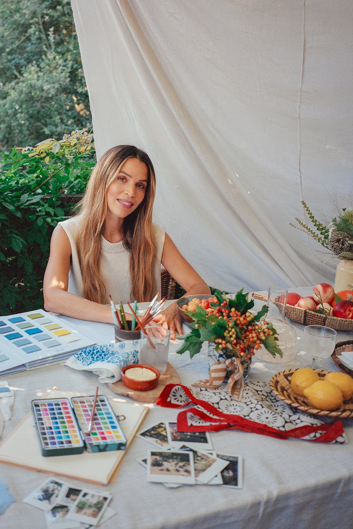 A woman sits at a table filled with art supplies, fruits, and plants, set against the lightweight woven cotton backdrop of the NOLA SCARF by Velvet by Graham & Spencer. The scene exudes creativity and tranquility under the open sky.-38895974842561