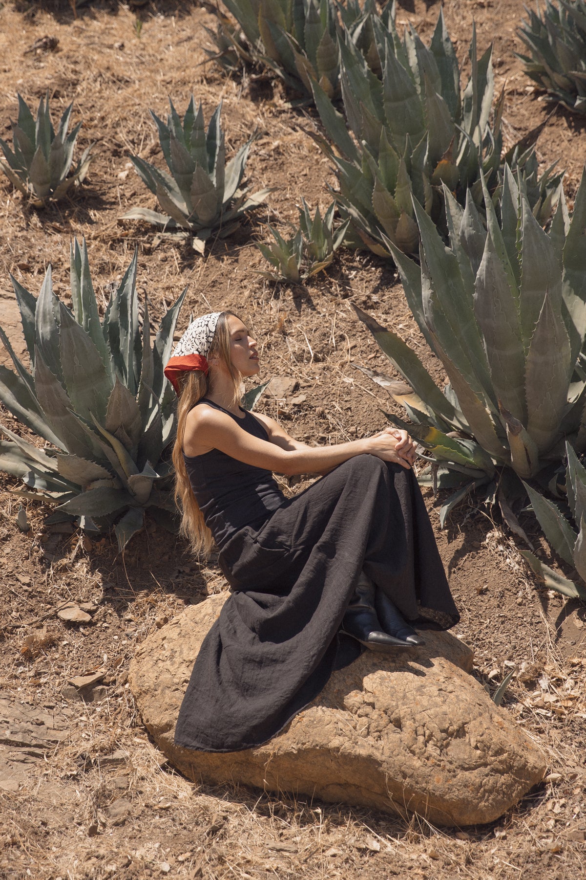 A woman with a red bandana sits on a rock among agave plants in a dry, grassy landscape, wearing the GREER TANK TOP by Velvet by Graham & Spencer and boots.-38819648864449