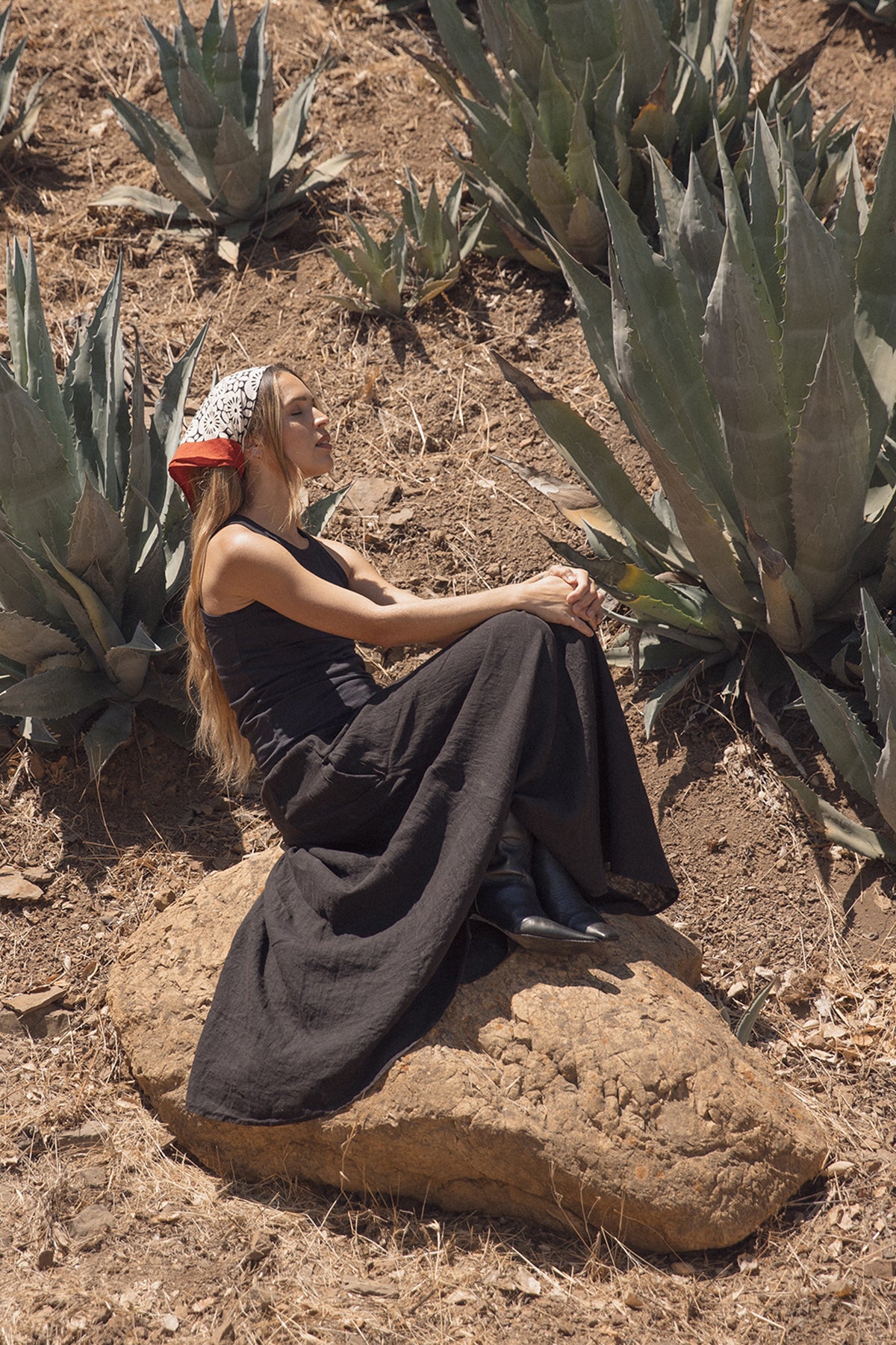 A woman wearing a black dress and the NOLA SCARF by Velvet by Graham & Spencer sits on a rock in the desert, surrounded by large agave plants.-38864719839425