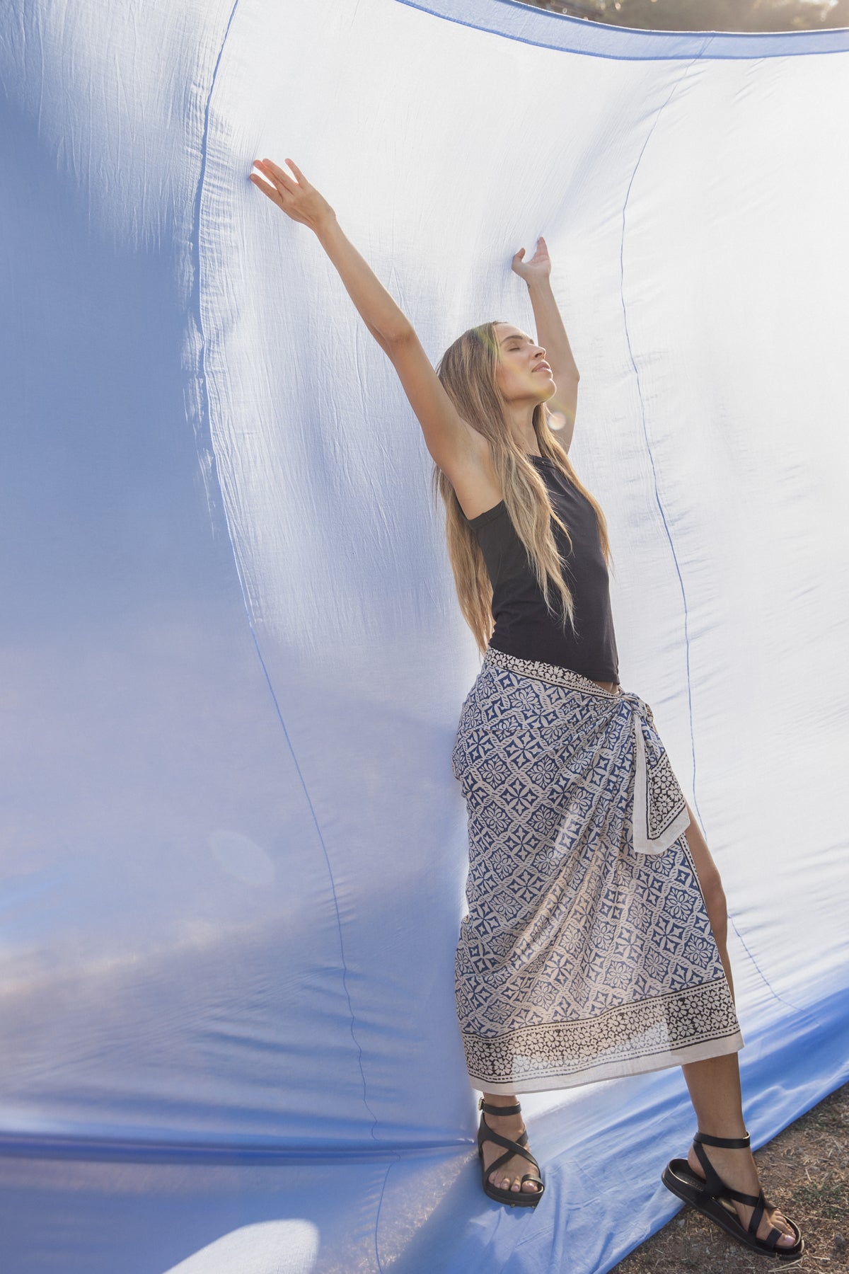 A person stands outside with arms raised against a large blue fabric, wearing the GREER TANK TOP by Velvet by Graham & Spencer, a patterned skirt, and sandals.-38819647455425