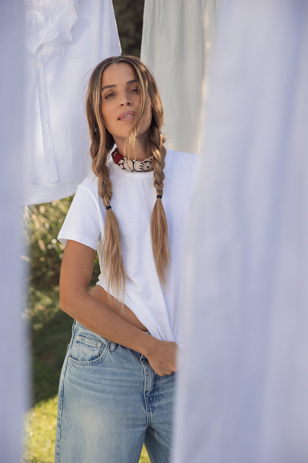 A person with long braids stands between hanging white sheets wearing a white t-shirt and jeans, embodying the versatile design spirit of the NOLA SCARF by Velvet by Graham & Spencer.-38890325082305