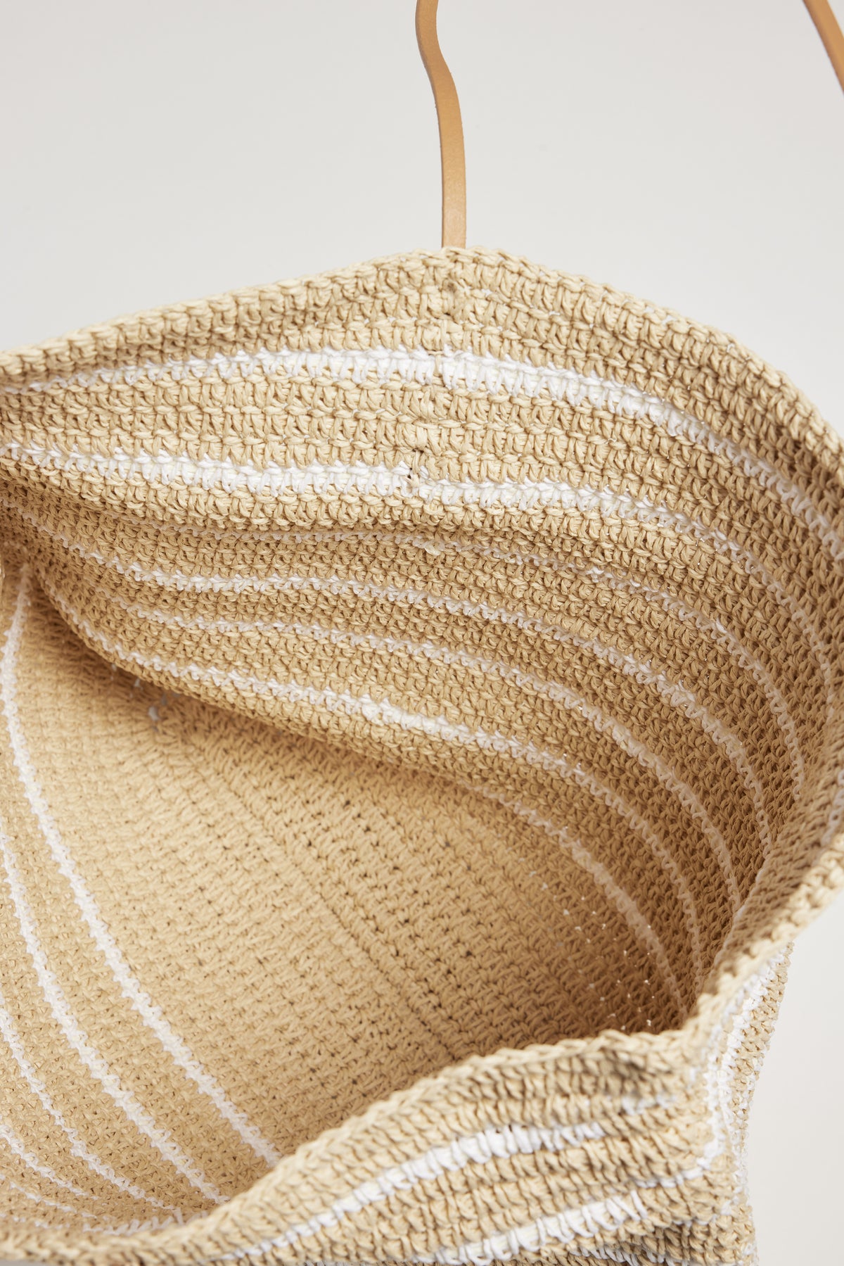   Close-up view of a versatile accessory, a Velvet by Graham & Spencer woven straw hat hanging on a hook against a plain background, highlighting its textured details and curved brim. 