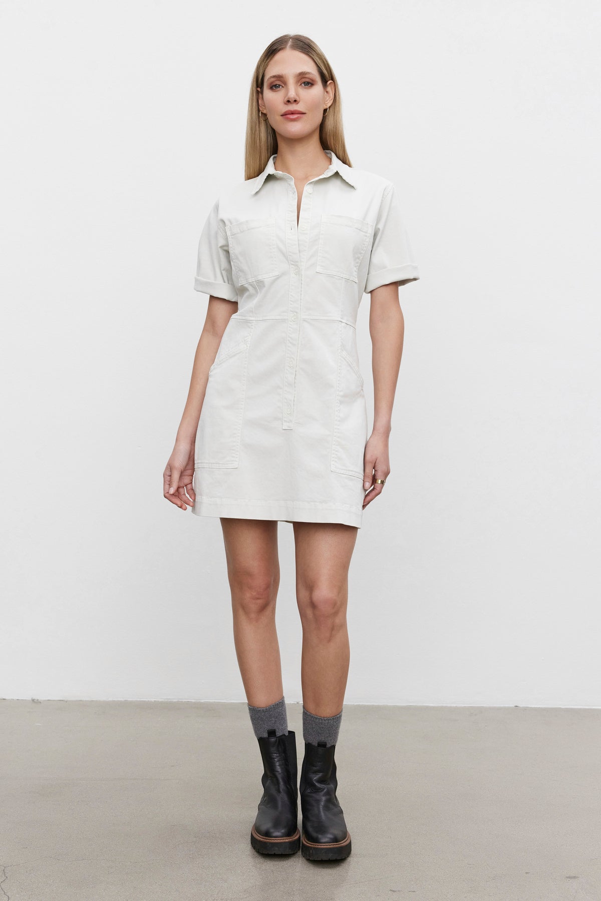   A woman stands against a plain backdrop wearing the CRISTINA DRESS from Velvet by Graham & Spencer, which features short sleeves, a button-down front, and a detachable belt. She pairs it with black ankle boots and gray socks. 