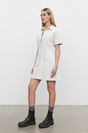 Person standing in profile view wearing the CRISTINA DRESS from Velvet by Graham & Spencer—a white short-sleeve shirt dress made of mid-weight cotton twill—and black boots with gray socks, on a concrete floor against a plain white background.