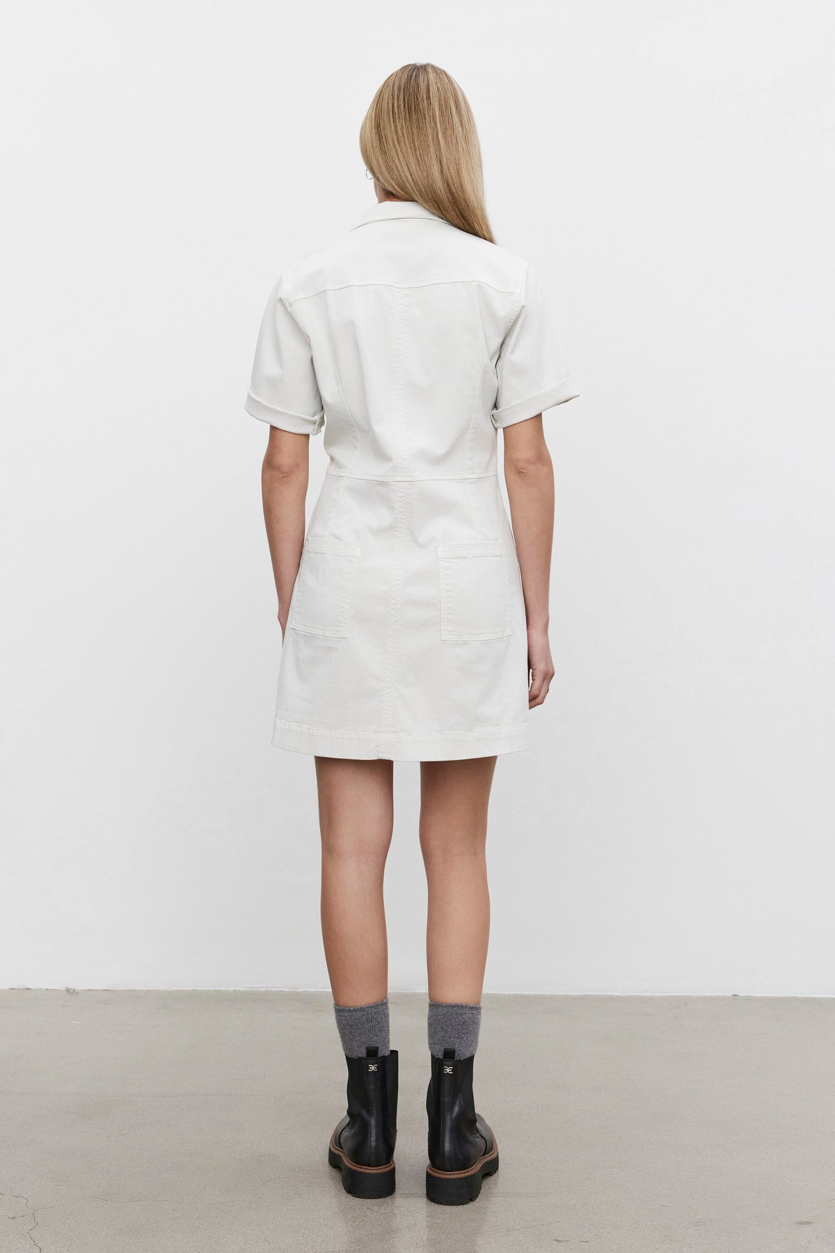   A person wearing the CRISTINA DRESS by Velvet by Graham & Spencer, a short-sleeved, knee-length white dress made of mid-weight cotton twill, stands facing away to showcase the back of the dress and is also wearing gray socks and black ankle boots. 