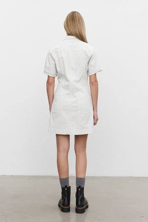 A person wearing the CRISTINA DRESS by Velvet by Graham & Spencer, a short-sleeved, knee-length white dress made of mid-weight cotton twill, stands facing away to showcase the back of the dress and is also wearing gray socks and black ankle boots.