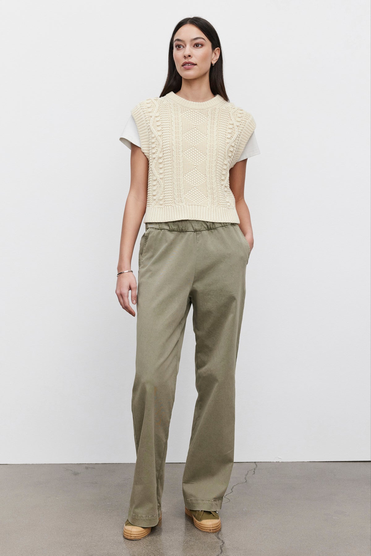 A person stands against a plain white background, wearing a beige knit vest over a white shirt and the NAYA PANT from Velvet by Graham & Spencer, crafted from olive green stretch cotton twill, paired with brown shoes.-38428777349313