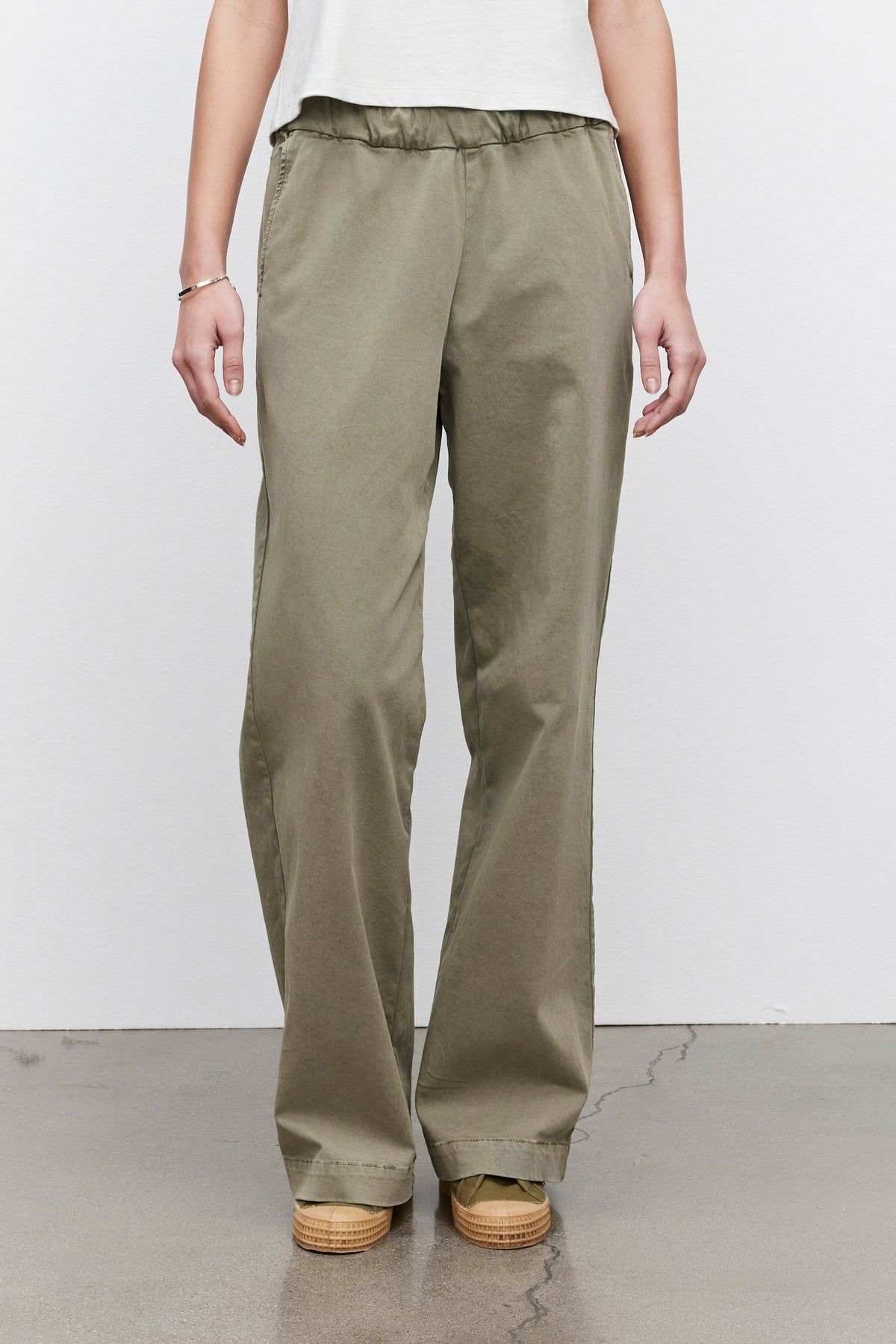   A person wearing the NAYA PANT in olive green from Velvet by Graham & Spencer and beige shoes stands against a plain white background with a concrete floor. The pants, made from stretch cotton twill, offer a comfortable fit. 