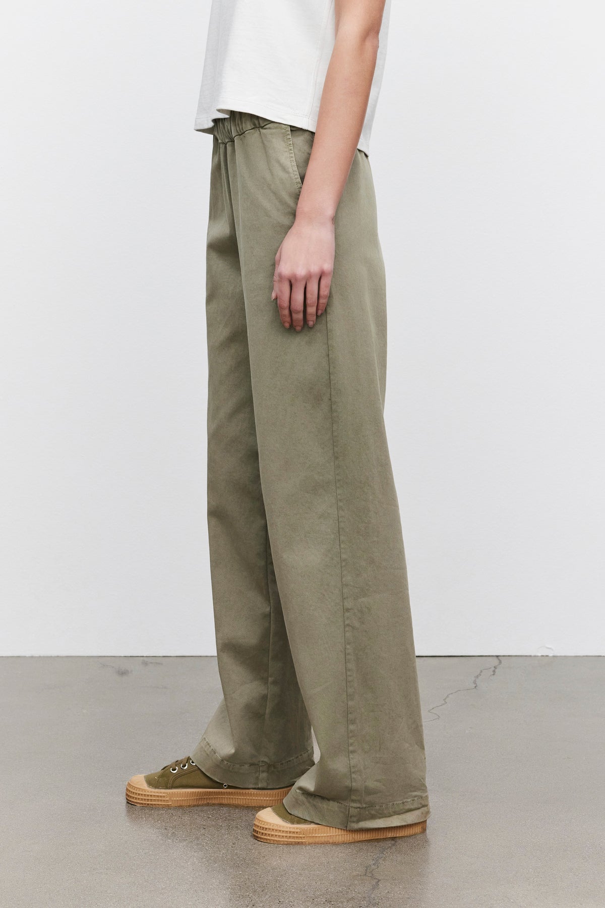   Person wearing the NAYA PANT by Velvet by Graham & Spencer in green and green sneakers, standing on a smooth floor with a white wall background. 