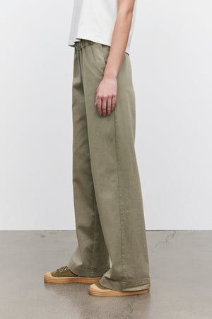 Person wearing the NAYA PANT by Velvet by Graham & Spencer in green and green sneakers, standing on a smooth floor with a white wall background.