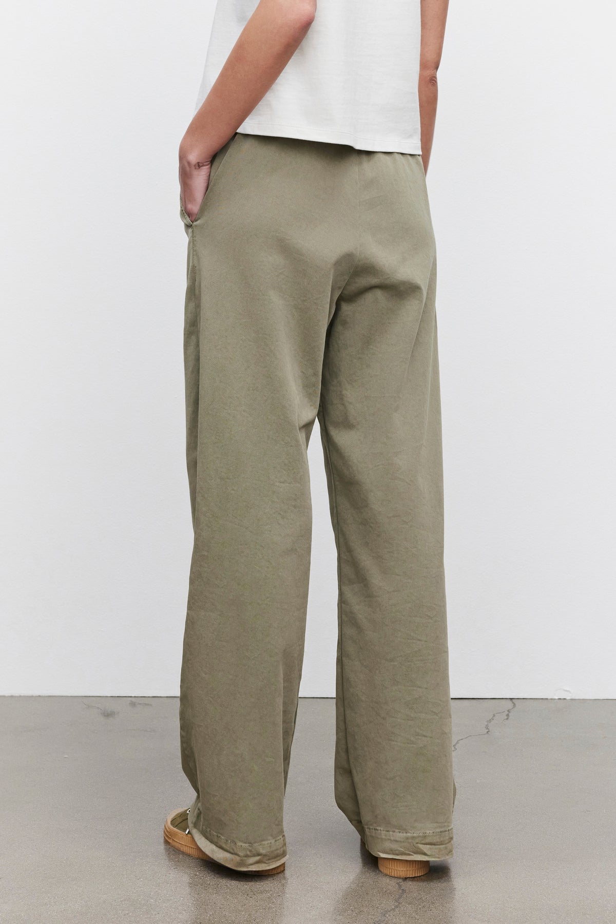   An individual wearing Velvet by Graham & Spencer's NAYA PANT in olive green, made from stretch cotton twill, paired with a white short-sleeve top, is standing with hands in pockets and facing away. 