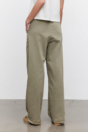 An individual wearing Velvet by Graham & Spencer's NAYA PANT in olive green, made from stretch cotton twill, paired with a white short-sleeve top, is standing with hands in pockets and facing away.