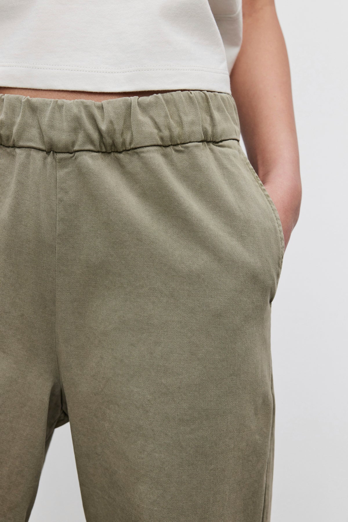   Close-up of a person wearing the NAYA PANT from Velvet by Graham & Spencer, featuring wide-leg design in olive green stretch cotton twill with a comfortable fit, one hand resting in the pocket, paired with a white shirt. 