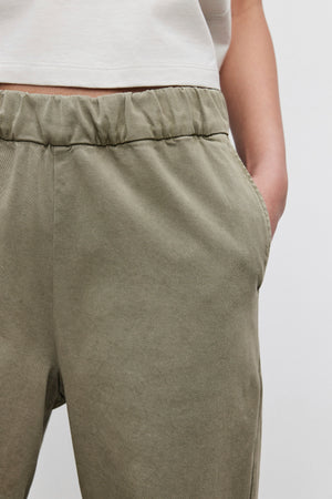 Close-up of a person wearing the NAYA PANT from Velvet by Graham & Spencer, featuring wide-leg design in olive green stretch cotton twill with a comfortable fit, one hand resting in the pocket, paired with a white shirt.