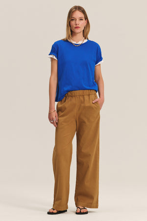A woman wears Velvet by Graham & Spencer's NAYA PANT—stretch cotton twill wide-leg pants with an elastic waistband—paired with a blue tee over a white shirt, black sandals, a necklace, and has one hand in her pocket.