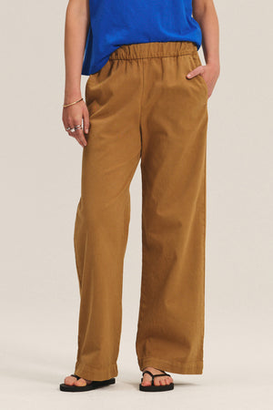 A person stands against a plain background wearing the Velvet by Graham & Spencer NAYA PANT—wide-leg brown stretch cotton twill with an elastic waistband—paired with a blue shirt, black sandals, and silver rings, one hand in their pocket.