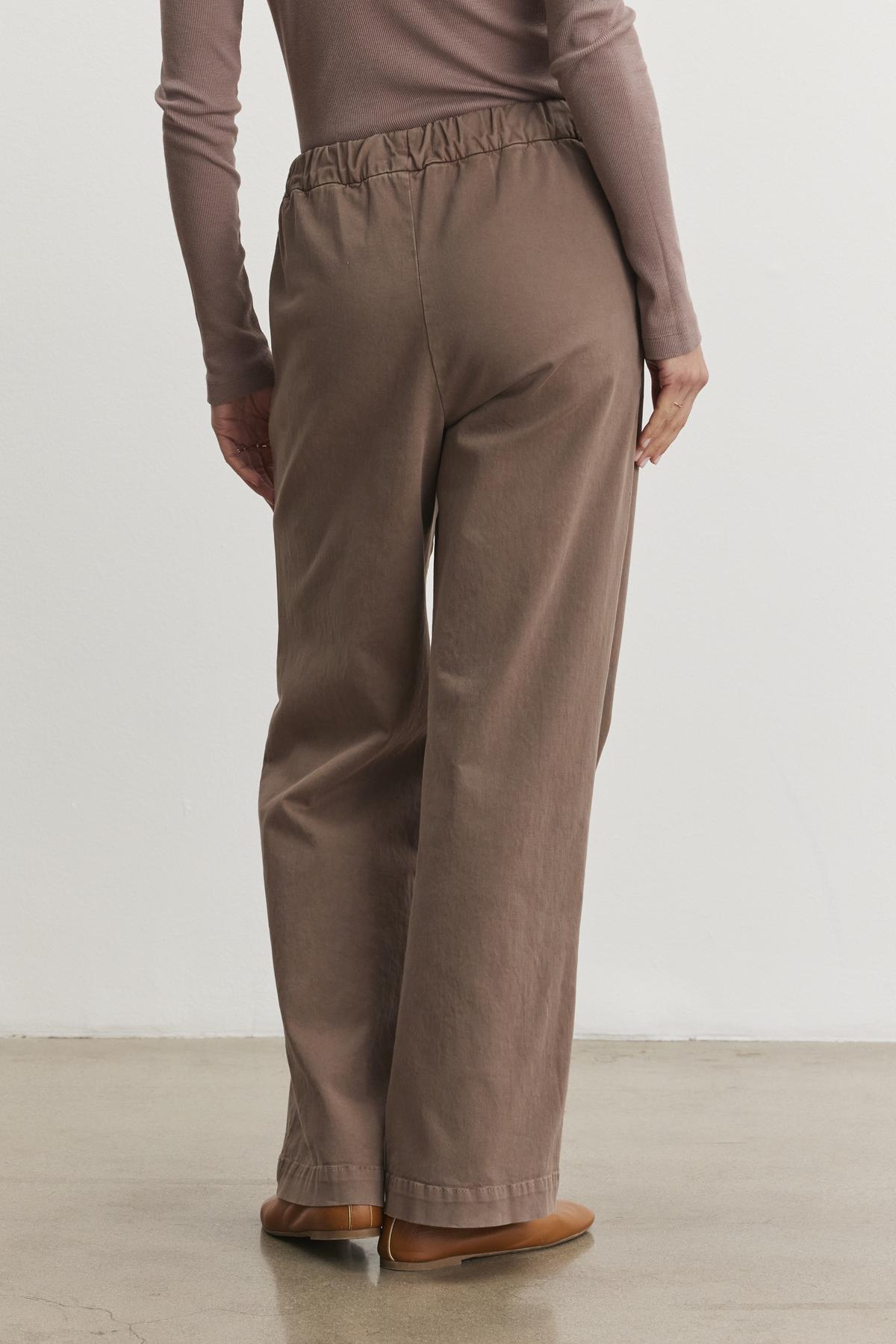   The individual is dressed in a long-sleeve brown shirt paired with Velvet by Graham & Spencer's NAYA PANT, featuring a wide-leg design made from stretch cotton twill. This ensemble is completed with flat shoes as they stand on a concrete floor, seen from behind. 