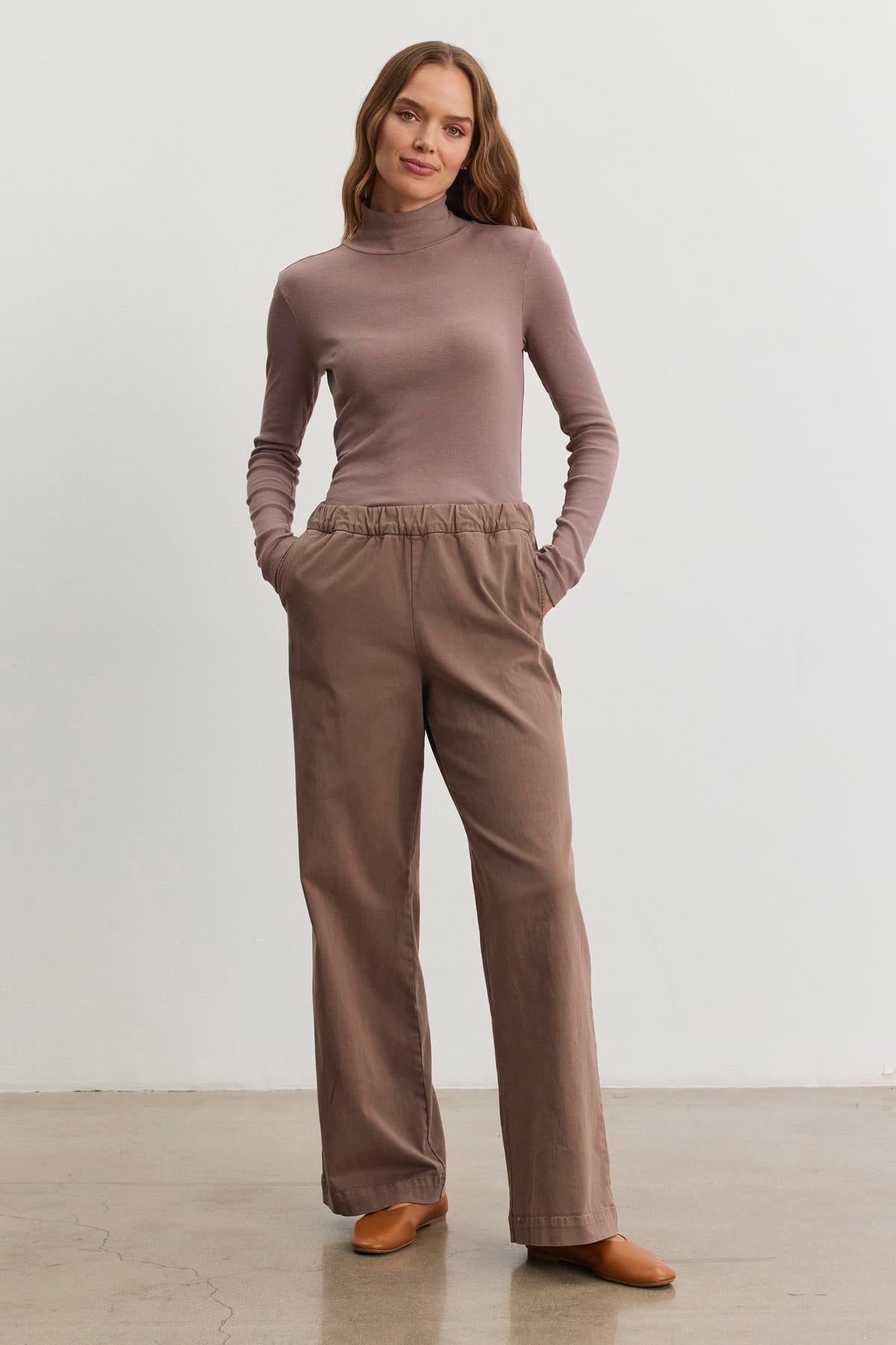   A woman in a taupe turtleneck and the NAYA PANT by Velvet by Graham & Spencer, featuring brown wide-leg design, stands against a plain background with hands in pockets and a neutral expression, her outfit showcasing stretch cotton twill for added comfort. 