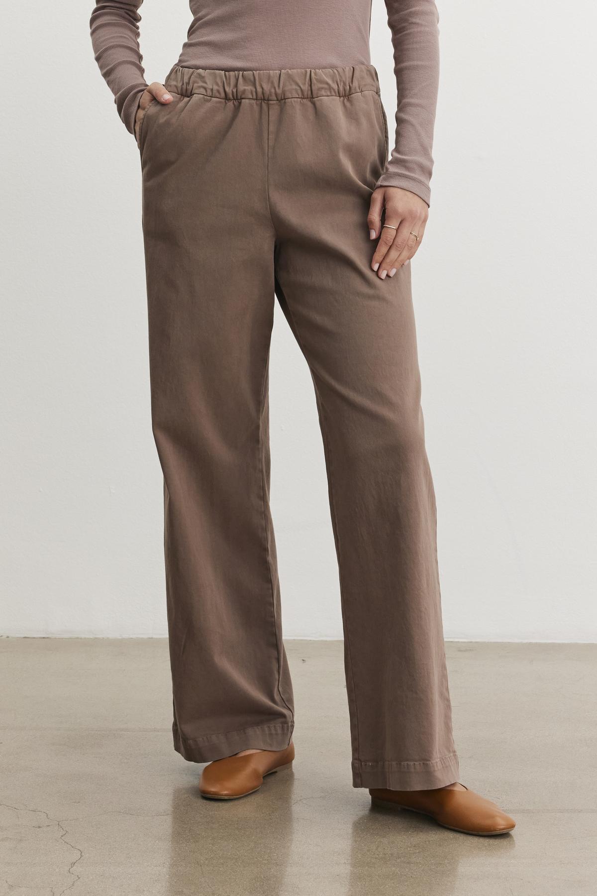   A person wearing a long-sleeve top and the NAYA PANT by Velvet by Graham & Spencer, featuring brown stretch cotton twill wide-leg pants with hands in pockets, standing on a polished floor. 