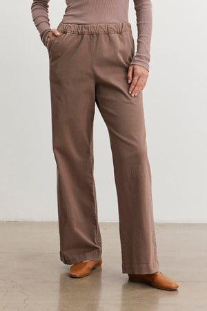 A person wearing a long-sleeve top and the NAYA PANT by Velvet by Graham & Spencer, featuring brown stretch cotton twill wide-leg pants with hands in pockets, standing on a polished floor.