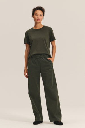 A person stands against a plain background wearing the Velvet by Graham & Spencer NAYA PANT, styled with a loose dark green t-shirt, black shoes, and one hand in their pocket.