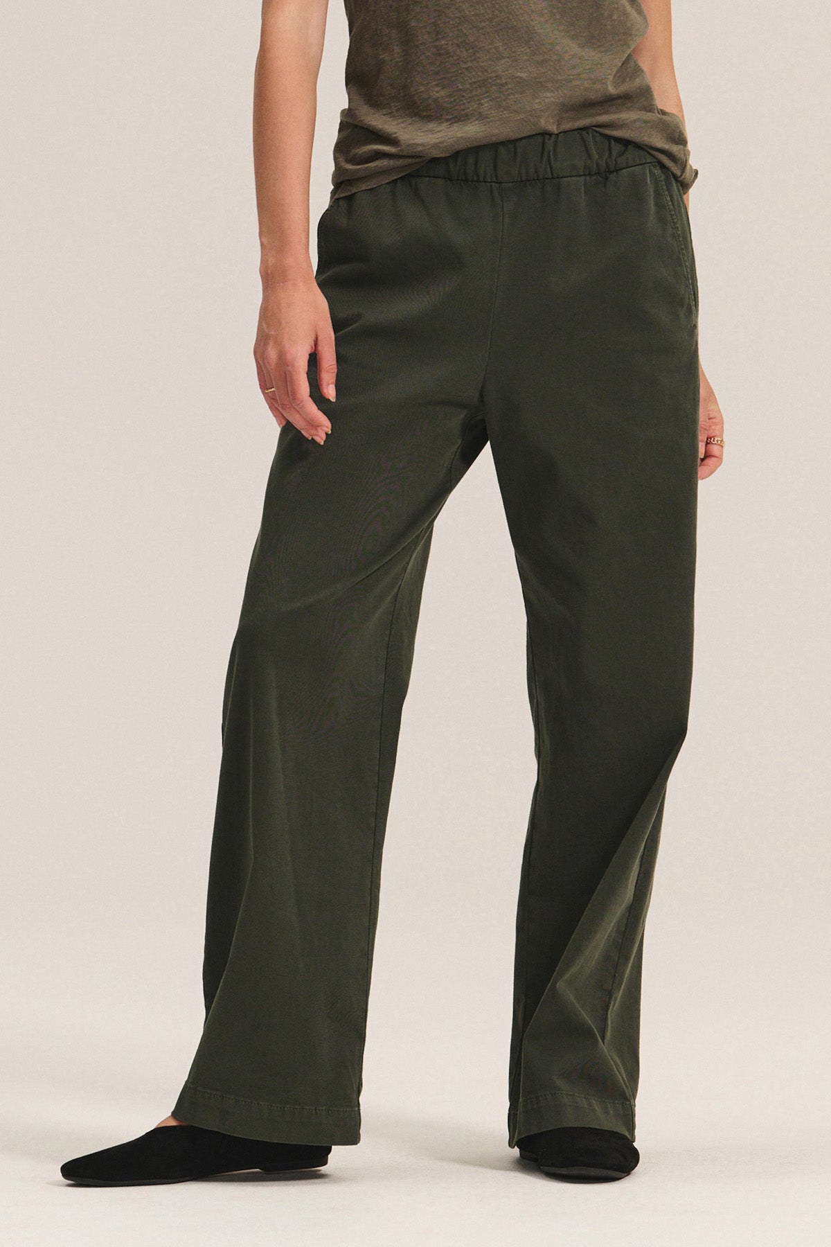 A person wears the NAYA PANT by Velvet by Graham & Spencer—wide-leg, dark green stretch cotton twill pants with an elastic waistband—paired with a brown top and black shoes, standing against a light background.-40248446681281