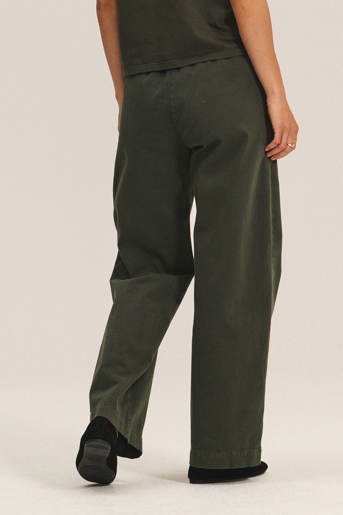 A person faces away wearing the Velvet by Graham & Spencer NAYA PANT in dark green stretch cotton twill with a wide leg and elastic waistband, paired with a matching top and black shoes, set against a neutral background.