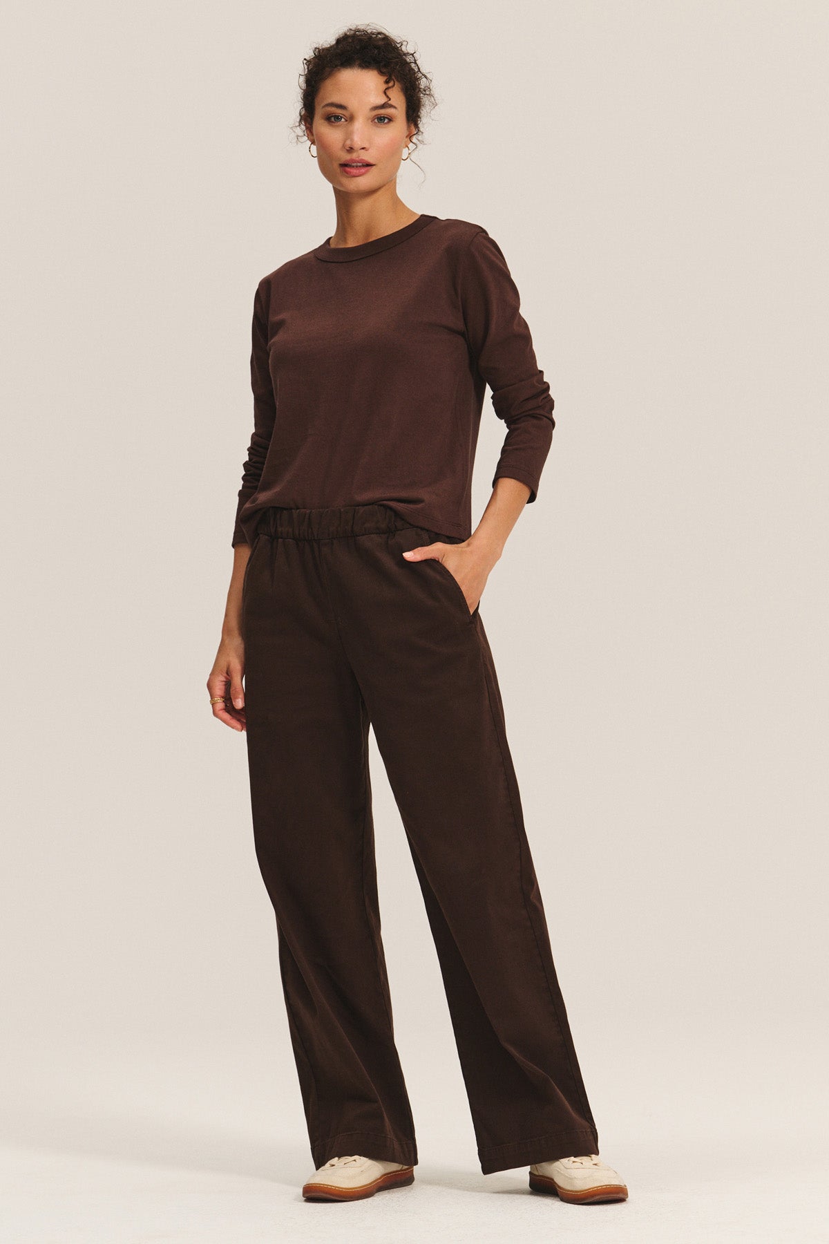 A woman wears the NAYA PANT by Velvet by Graham & Spencer—stretch cotton twill wide-leg pants with an elastic waistband—paired with a brown long-sleeve top and white sneakers, standing against a light plain background.-40263665418433