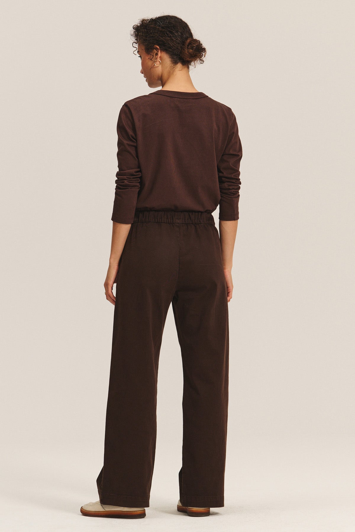A person stands against a plain light background, wearing the NAYA PANT by Velvet by Graham & Spencer with an elastic waistband, paired with a long-sleeve brown shirt and flat shoes, facing away from the camera.-40263665385665