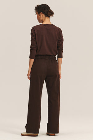 A person stands against a plain light background, wearing the NAYA PANT by Velvet by Graham & Spencer with an elastic waistband, paired with a long-sleeve brown shirt and flat shoes, facing away from the camera.