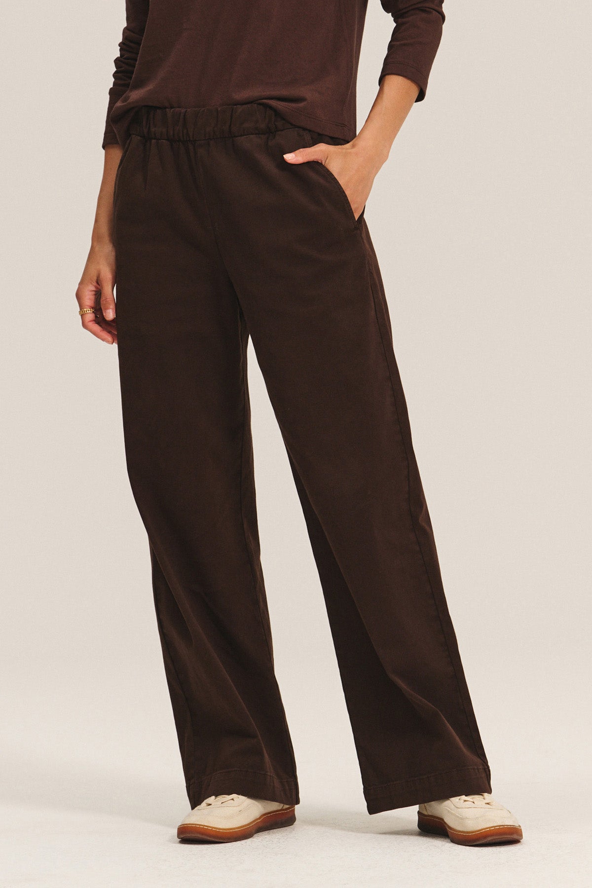 Model wears the NAYA PANT by Velvet by Graham & Spencer—loose, wide-leg dark brown pants with an elastic waistband—paired with a matching long-sleeve top and cream-colored shoes, standing with one hand in a pocket.-40263659782337