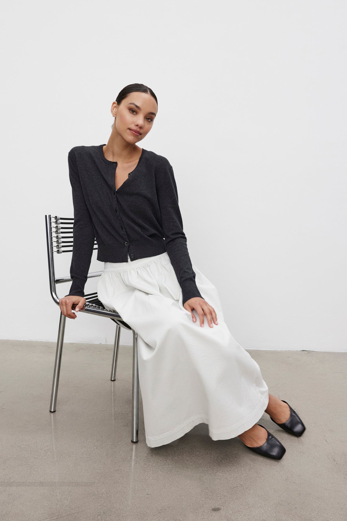   A person with dark hair wearing a VIOLETTE CARDIGAN by Velvet by Graham & Spencer and a long white skirt sits on a metal chair against a plain white background. 