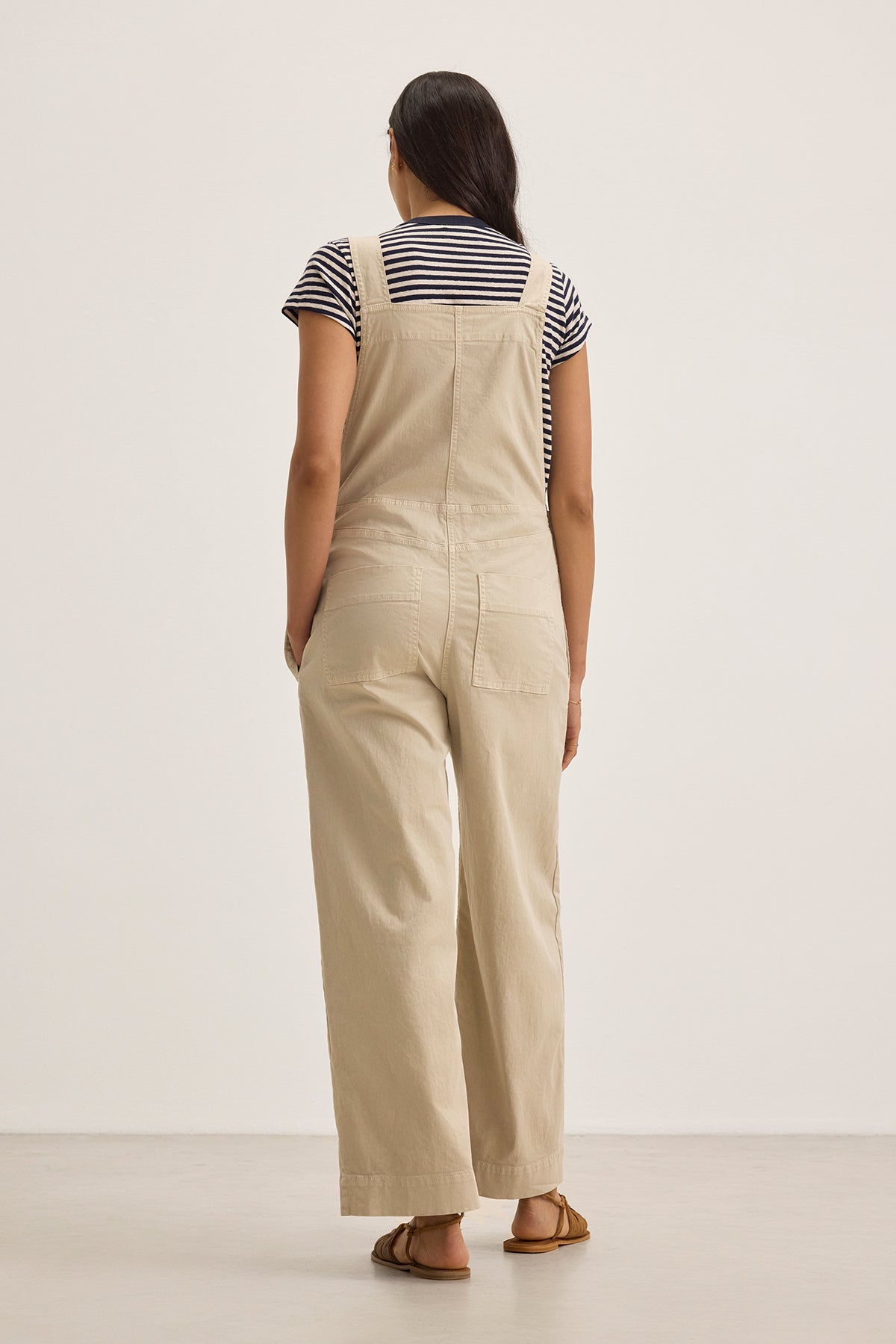 A person wearing the Velvet by Graham & Spencer DREA JUMPSUIT, crafted from stretch cotton twill in a wide-leg silhouette with adjustable straps for comfort and style, stands showcasing its beige design alongside a black and white striped t-shirt against a plain backdrop.-38662928466113