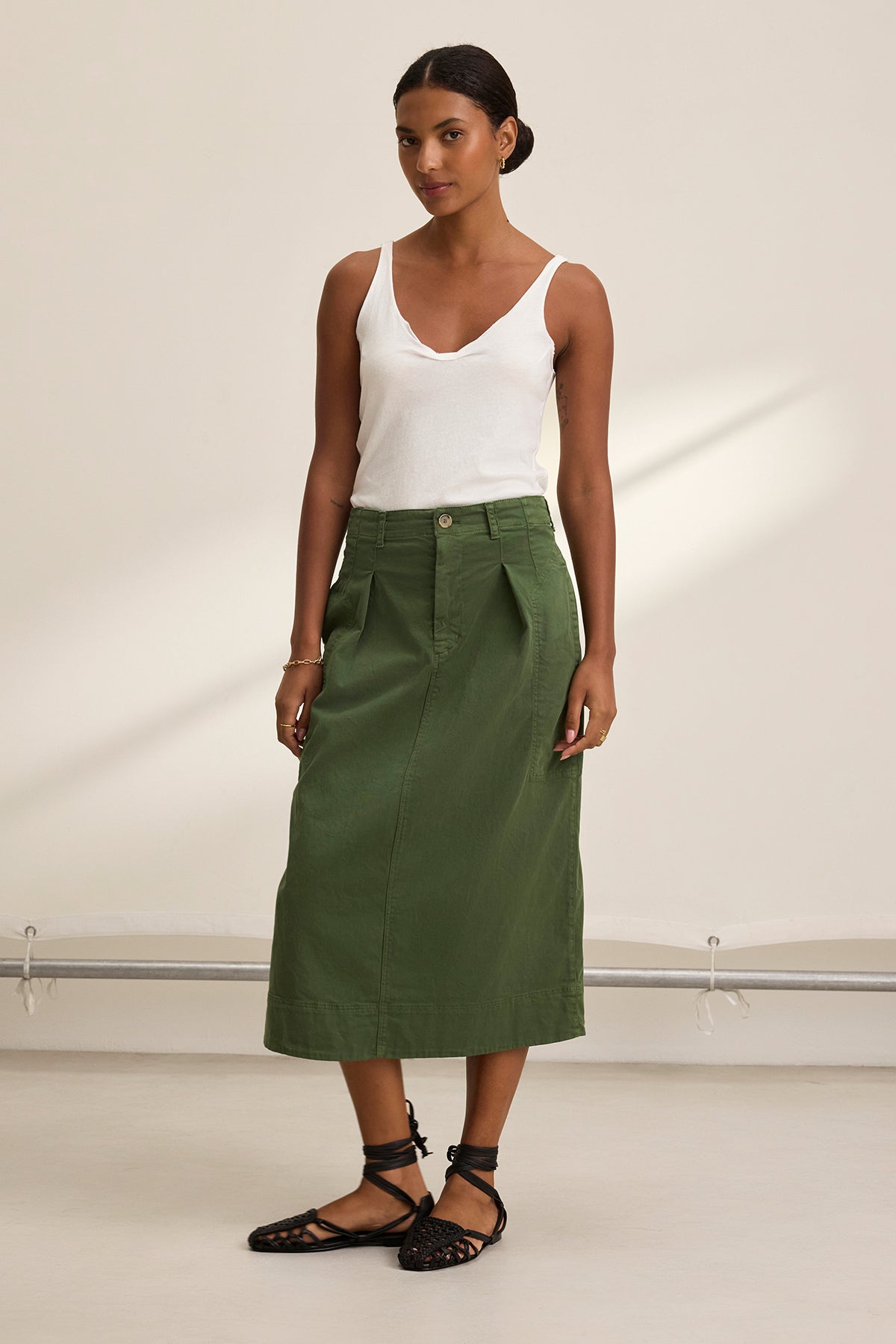 Standing against a neutral background, the person wears a white sleeveless top, black shoes, and the LILIAN SKIRT by Velvet by Graham & Spencer, a green A-line skirt made from stretch cotton twill that blends style and comfort for any occasion.-38662723043521