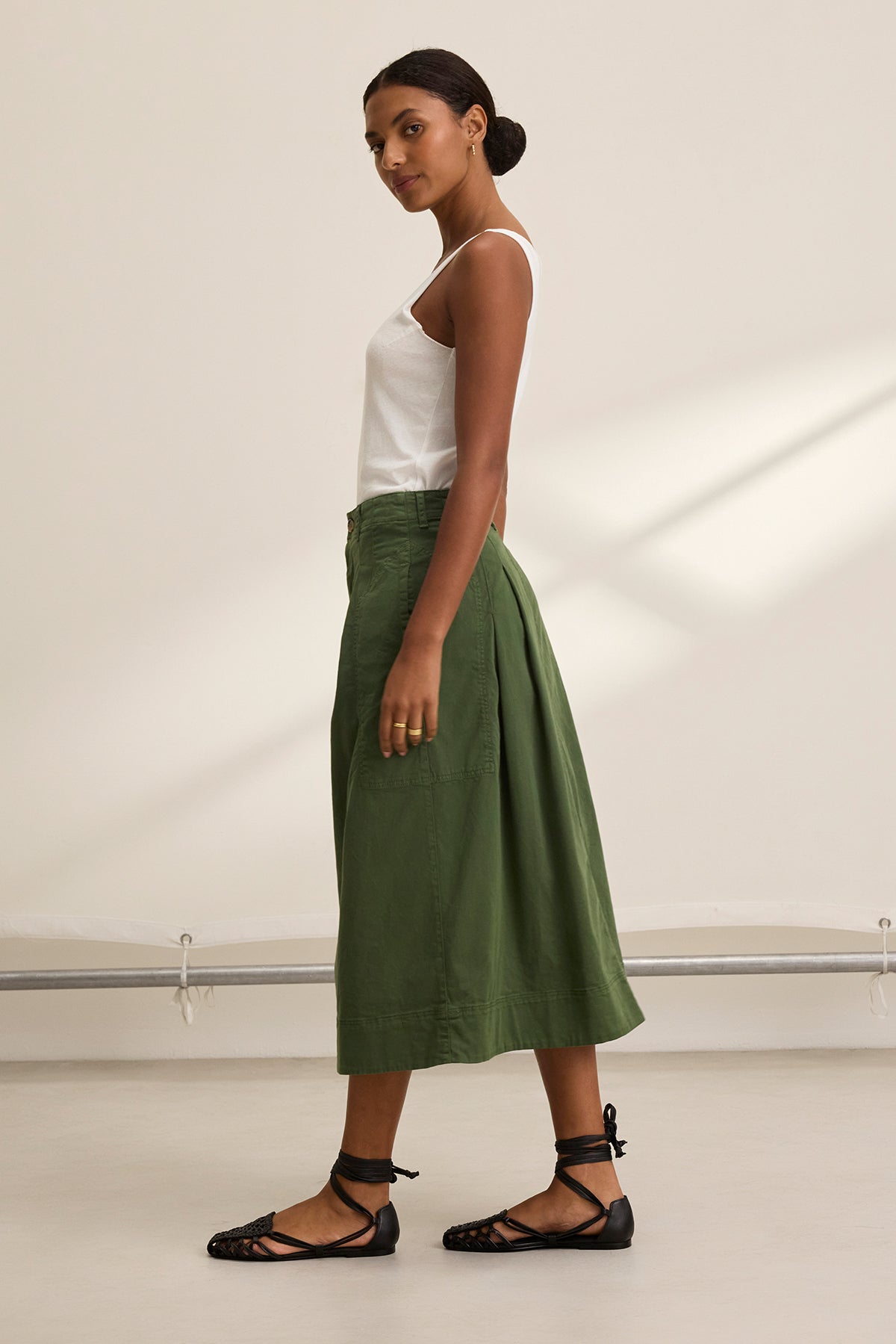 A woman showcases the Velvet by Graham & Spencer's LILIAN SKIRT, a green stretch cotton twill A-line skirt, paired with a sleeveless white top and black lace-up sandals. This versatile piece effortlessly transitions from casual day outings to evening events.-38662723076289