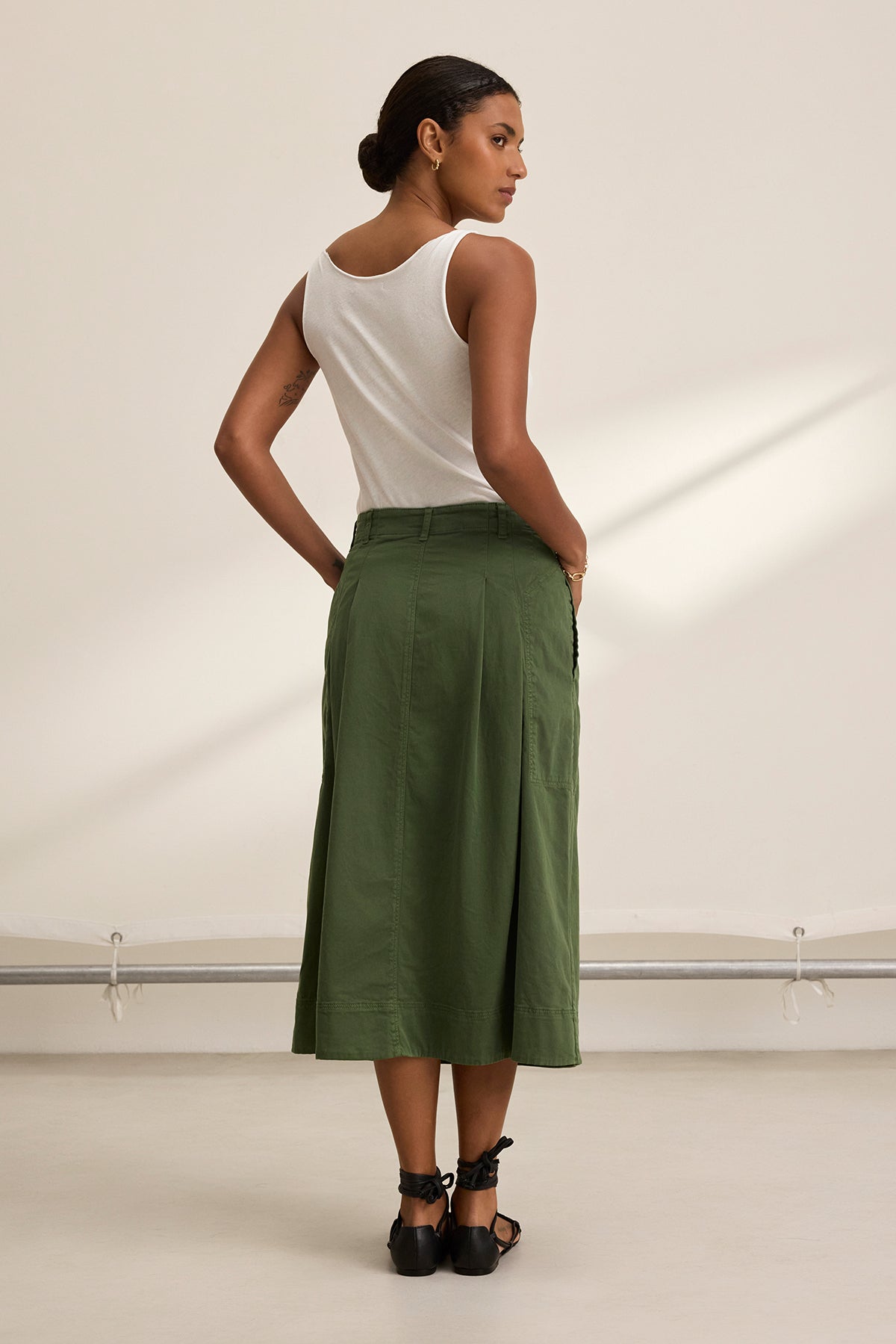 A person facing away, donning a white sleeveless top, an olive green LILIAN SKIRT by Velvet by Graham & Spencer made from stretch cotton twill, and black sandals stands in a neutral-toned room.-38662723109057