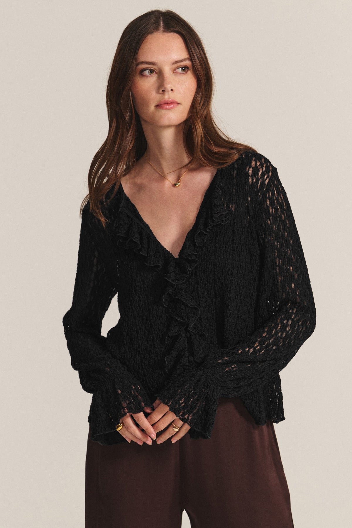 A woman wears the NATALIE TOP by Velvet by Graham & Spencer, a textured black long-sleeve cotton lace top with ruffle details, paired with brown pants and standing against a plain background.-40681825960129