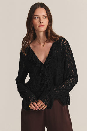 A woman wears the NATALIE TOP by Velvet by Graham & Spencer, a textured black long-sleeve cotton lace top with ruffle details, paired with brown pants and standing against a plain background.