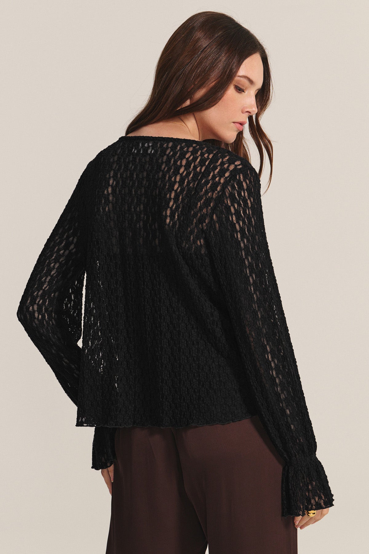 A woman with long brown hair wears the Velvet by Graham & Spencer NATALIE TOP, a black, long-sleeve, semi-sheer blouse with subtle ruffle details, paired with brown pants as she stands slightly turned away from the camera.-40681825927361