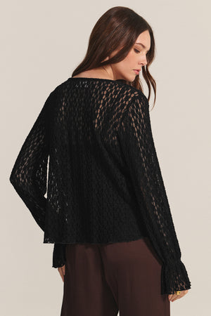 A woman with long brown hair wears the Velvet by Graham & Spencer NATALIE TOP, a black, long-sleeve, semi-sheer blouse with subtle ruffle details, paired with brown pants as she stands slightly turned away from the camera.