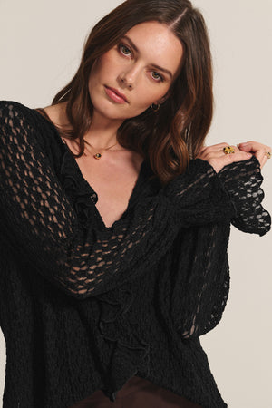 A woman with wavy brown hair wears the NATALIE TOP by Velvet by Graham & Spencer—a black cotton lace top with ruffle detailing and textured long sleeves—standing against a plain light background.