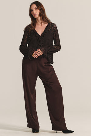 A woman wears the NATALIE TOP by Velvet by Graham & Spencer—a black cotton lace top with ruffles and a detachable cami—paired with loose dark brown satin pants and black pointed-toe shoes.