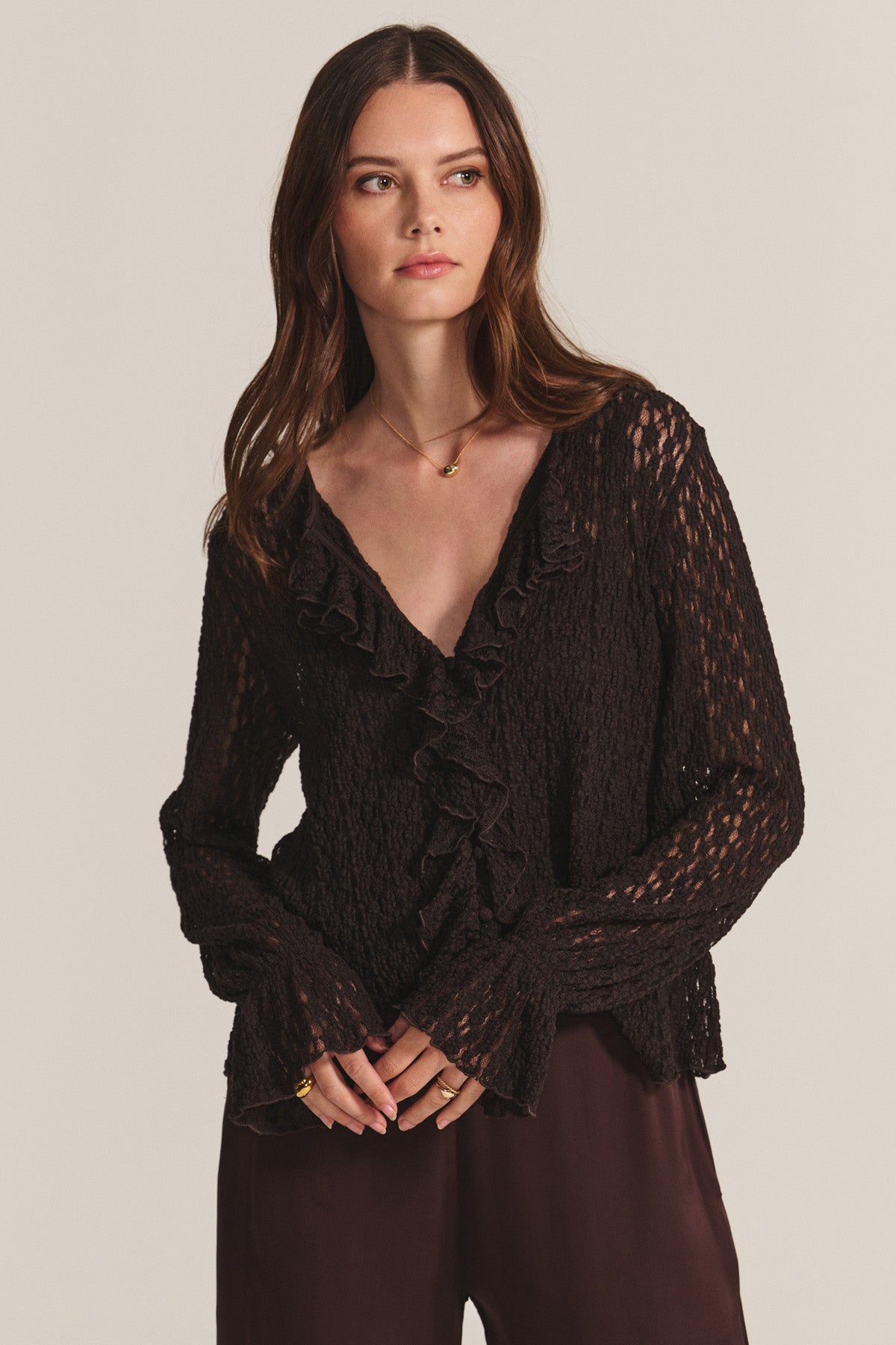 A woman stands against a plain background wearing the NATALIE TOP by Velvet by Graham & Spencer—a textured, long-sleeve black blouse with ruffle detailing at the neckline—paired with dark brown pants.