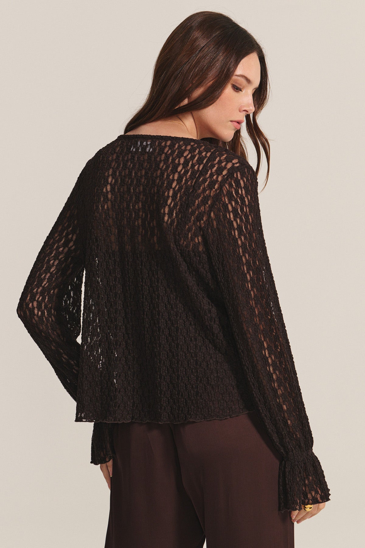 A woman with long brown hair is shown from the back against a neutral background, wearing the Velvet by Graham & Spencer NATALIE TOP—a loose, long-sleeve cotton lace top—paired with dark pants.